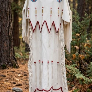 Puede incluir: Un vestido de cuero color hueso con largos flecos en las mangas y los laterales. La prenda está decorada con parches circulares de cuentas rojas y azules, ribetes de cuentas rojas en el cuello y el dobladillo, y filas de cuentas cilíndricas.