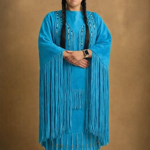 May include: A turquoise dress with long fringe and beaded accents. The dress has long sleeves and a matching skirt. A single feather is placed in the hair. The person is wearing a smartwatch.