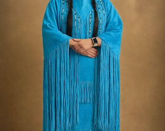 May include: A turquoise dress with long fringe and beaded accents. The dress has long sleeves and a matching skirt. A single feather is placed in the hair. The person is wearing a smartwatch.