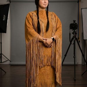 May include: A woman wearing a tan suede dress with long fringe and a matching shawl. She has two long braids, a feather in her hair, and a small tattoo on her face. The dress is a traditional Native American style.