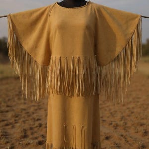 Puede incluir: Un vestido de dos piezas de color beige con una capa y una falda con flecos. La capa tiene flecos largos en los bordes y la falda tiene flecos en el dobladillo. El vestido se exhibe en un maniquí negro sobre un fondo exterior borroso.