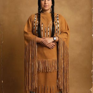 Puede incluir: Un vestido color marrón con mangas largas y dobladillo con flecos. El vestido está adornado con detalles de cuentas blancas, negras y turquesas. La mujer de la imagen tiene largas trenzas negras y lleva un reloj inteligente.