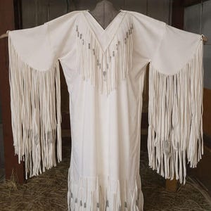 Könnte beinhalten: Cremefarbenes Kleid mit langen Ärmeln und V-Ausschnitt. Das Kleid hat lange Fransen an den Ärmeln, am Ausschnitt und am Saum. Das Kleid hängt an einem Holzkleiderbügel.