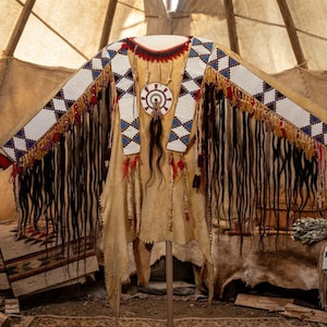 May include: A tan leather shirt featuring long dark fringe, intricate white, blue, and red beadwork in diamond patterns along the sleeves, and a circular beaded medallion on the chest with a central sunburst design and hanging dark hair tassels.