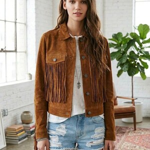 Pode incluir: Casaco de camurça castanho com franjas nas mangas e bolsos no peito. O casaco é usado sobre uma camiseta branca e calções de ganga azul clara. Um colar de prata completa o look.