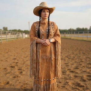 Puede incluir: Una mujer con un vestido de ante color beige con flecos y adornos de cuentas, y un sombrero de vaquero. El vestido tiene mangas largas y cuello alto. Está de pie en un ruedo de tierra, con una valla al fondo.