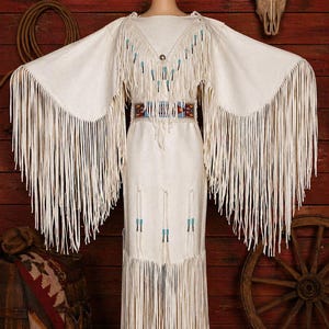May include: Cream-colored fringed dress with long sleeves and a decorative beaded yoke. The dress features a detailed belt and fringe along the hemline. The dress is displayed on a mannequin against a rustic wooden background.