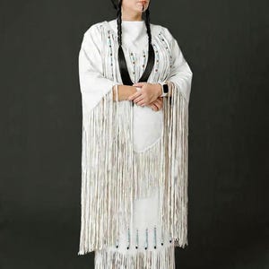 May include: A woman in a white fringed dress and shawl, adorned with beaded accents. She wears a feather in her hair and has black braids. The dress features intricate fringe detailing and is paired with embroidered moccasins, set against a dark background.