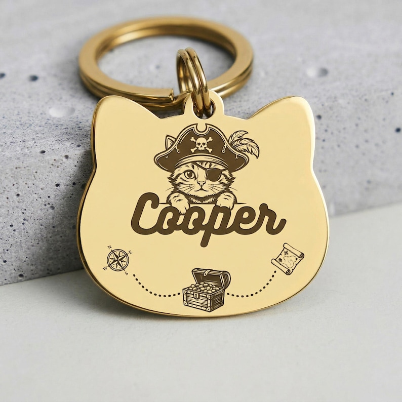 Peut inclure: Une m&eacute;daille d'identification pour animal en m&eacute;tal dor&eacute; en forme de chat, grav&eacute;e d'un chaton portant un chapeau de pirate et un cache-&oelig;il. Le nom Cooper est grav&eacute; en lettres cursives sous le chaton, avec une boussole, un coffre et une carte.