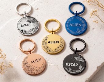 Peut inclure: Plusieurs porte-clés ronds en métal avec divers motifs et le mot "ALIEN" ou un nom. Les porte-clés présentent des images d'OVNI, de paysages désertiques et de motifs floraux. Les couleurs incluent l'argent, l'or, l'or rose, le bleu et le noir.