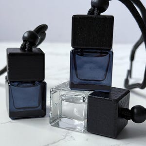 May include: Several small, square glass bottles with black wooden tops and black cords. Two bottles have a dark blue liquid inside, while one is clear. The bottles are arranged on a white marble surface.