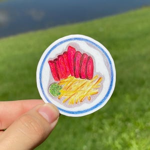 May include: A circular sticker featuring a colored pencil drawing of a white plate with a blue rim. The plate contains several slices of bright pink tuna sashimi, a pile of thin yellow french fries, and a small scoop of textured green wasabi.