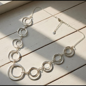 Puede incluir: Un collar plateado con una serie de círculos irregulares interconectados. El collar tiene una cadena fina y un cierre ajustable. El diseño es moderno y minimalista, adecuado para el uso diario o ocasiones especiales.