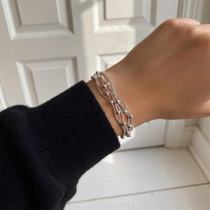 May include: A silver bracelet featuring multiple strands and small, round beads. The bracelet is worn on a wrist, with a black long-sleeve top in the background. The bracelet is a delicate piece of jewellery.
