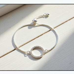 May include: A silver bracelet with a circular pendant. The bracelet has a fine chain and a round pendant set with small, sparkling stones. The adjustable clasp allows for a custom fit. The jewellery is displayed on a white surface.