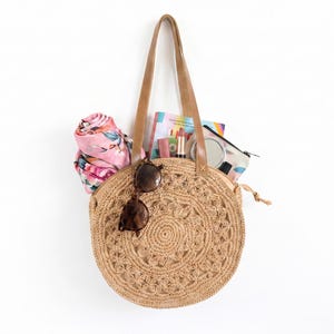 May include: A round, woven straw handbag with brown leather handles, filled with a pink floral scarf, sunglasses, and makeup. The bag has a decorative floral pattern and is hanging against a white background.
