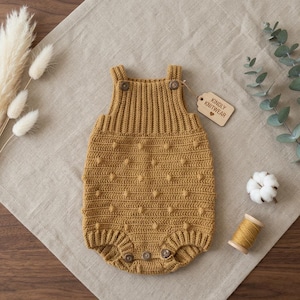 以下が含まれることがあります： マスタードイエローのニットベビーロンパース。木製のボタン留めと、テクスチャード加工されたドット柄が特徴です。「KINDLY KNITWEAR」と書かれたタグが付いています。ニュートラルなリネンの上に置かれています。