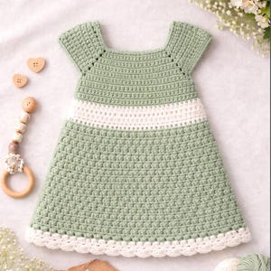 May include: A handmade crochet baby dress in a soft green colour with a white stripe and trim. The dress features a square neckline and short sleeves. A wooden baby toy and heart-shaped buttons are next to the dress.