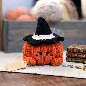 May include: A knitted orange pumpkin plush toy wearing a black witch hat with a white band. The pumpkin has black bead eyes and embroidered facial features. Beside it are three miniature books titled Autumnal Mysteries, Knit Spells, and The Tiny Witch.