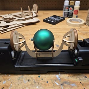May include: A black motorised rotary tool with a matte green spherical ornament mounted on a light-coloured wooden cradle. The device features a base with a length track and side supports, designed to rotate the sphere for crafting or painting tasks.