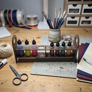 May include: A dark wood craft organiser stand featuring two rows of circular cutouts holding seven small plastic squeeze bottles filled with various coloured liquids. The stand has arched side panels and sits on a workspace with art supplies.