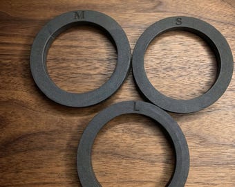 May include: Three circular, dark gray rings with a matte finish. Each ring features a single embossed letter on the top surface: one is marked with an M, one with an S, and one with an L. The rings are arranged in a triangular formation.