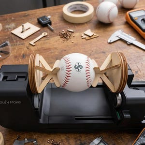 May include: A white baseball with red stitching held horizontally by two light-colored wooden supports, mounted on a black Eufy Make device. The baseball features a black logo with the text 00 and other small markings on its surface.