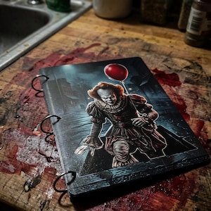 May include: A three-ring binder featuring a graphic illustration of a clown character with a white face, red hair, and a ruffled collar, emerging from a dark sewer grate while holding a single, bright red balloon against a dark, rainy street.