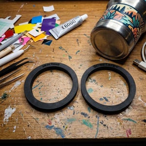 May include: Two black circular rings made of a matte material, each featuring embossed text. One ring is labeled 30 oz Small and the other is labeled 30 oz Large. These items are positioned on a work surface alongside crafting tools and a tumbler.