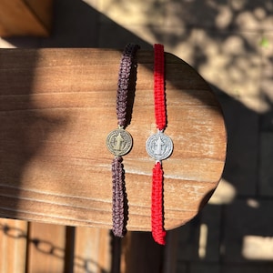 May include: Two macrame bracelets with Saint Benedict medals. One bracelet is dark brown, the other is red. The medals are round with a detailed design. The bracelets are displayed on a wooden surface.