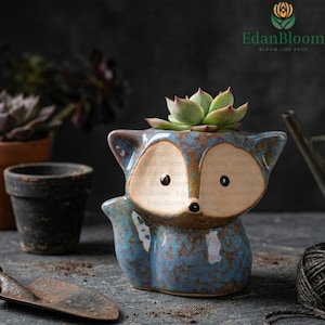 May include: A blue ceramic fox-shaped planter with a succulent plant. The planter has a distressed finish with brown accents. The fox has large, light-colored eyes and a black nose. Other potted plants and gardening tools are in the background.