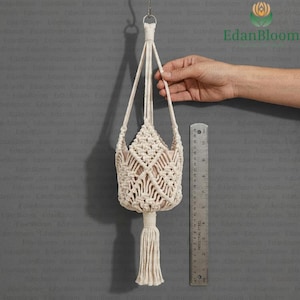 Pode incluir: Suporte de planta em macramé creme com um design de cesta tecida e uma longa borla. O suporte da planta é suspenso por um anel de metal e tem aproximadamente 25 cm de altura. Uma régua é colocada ao lado do suporte da planta.