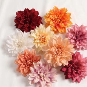 May include: A collection of artificial dahlia flowers in various colors, including deep red, orange, white, yellow, peach, and pink. The flowers are arranged on a white surface, showcasing their layered petal design. These decorative flowers are ideal for crafting and home decor.