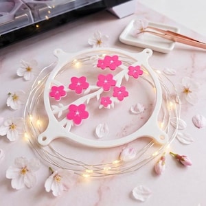 Può includere: Un elemento decorativo bianco e circolare con un ramo di fiori di ciliegio rosa. È circondato da una serie di luci LED bianche calde e petali di fiori di ciliegio sparsi. Un piccolo piatto bianco e delle pinzette sono sullo sfondo.