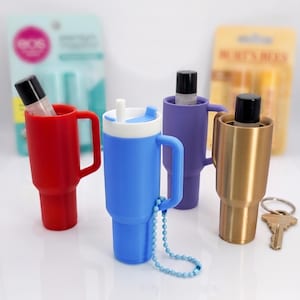 May include: Four colourful tumbler-shaped holders with handles, each holding a lip balm. The holders are red, blue, purple, and gold. The blue holder has a white lid and a blue beaded chain. A key and lip balm packaging are in the background.