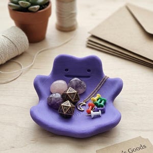 May include: A purple Ditto-shaped trinket dish holds amethyst stones, dice, a crescent moon necklace, and colorful push pins. A small tag reads "Gamer's Goods". A succulent in a terracotta pot and other craft supplies are in the background.