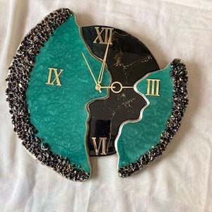 Puede incluir: Un reloj de pared decorativo con un diseño turquesa y negro. La esfera del reloj está dividida en secciones, con números romanos y manecillas doradas. Los bordes están adornados con adornos oscuros y texturizados.