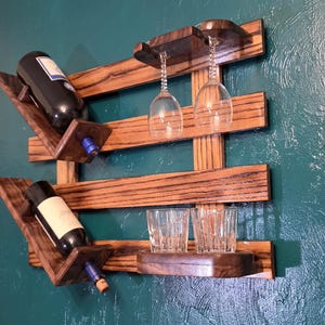 May include: A wall-mounted wooden wine rack featuring a dark brown wood grain finish. The rack holds two wine bottles at an angle, two hanging stemmed wine glasses, and two clear glass tumblers resting on a small wooden shelf on the bottom right.