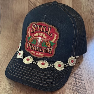 May include: A dark denim baseball cap featuring a red embroidered patch with the text STILL THE PROBLEM, a white steer skull, and orange roses. A silver-toned metal chain with circular concho-style accents and red stones drapes across the brim.