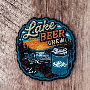 May include: A circular embroidered patch featuring a sunset over a lake with silhouettes of trees and a chair. The text reads Lake BEER CREW in cream and orange. A blue beverage can cooler with a white mountain graphic is in the foreground.