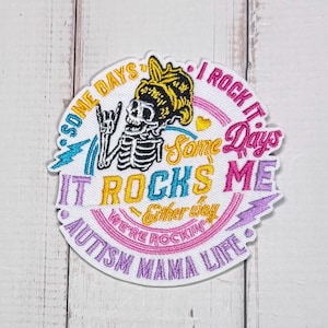 May include: A circular embroidered patch featuring a skeleton wearing a yellow headscarf and making a rock-and-roll hand gesture. The text reads: SOME DAYS I ROCK IT, Some Days IT ROCKS ME, Either way WE'RE ROCKIN, and AUTISM MAMA LIFE.