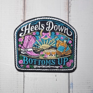 May include: A black embroidered patch with a white rope border featuring a pink cowboy boot, two amber-colored bottles, two shot glasses, and a golden sun. The text reads "HEELS DOWN" at the top and "BOTTOMS UP" at the bottom, surrounded by pink roses.