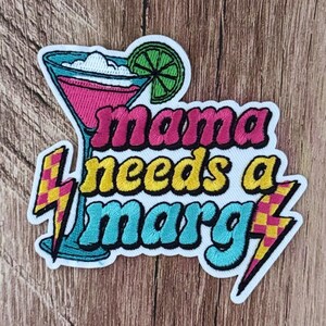 May include: An embroidered patch featuring a pink margarita in a glass with a green lime slice. The text reads mama needs a marg in pink, yellow, and blue letters. Two yellow and pink checkered lightning bolts flank the text on the sides.