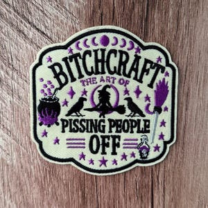 May include: An embroidered patch with a cream background and black border featuring the text BITCHCRAFT THE ART OF PISSING PEOPLE OFF. The design includes a black cauldron with purple bubbles, two black ravens, a witch on a broom, and a purple broom.