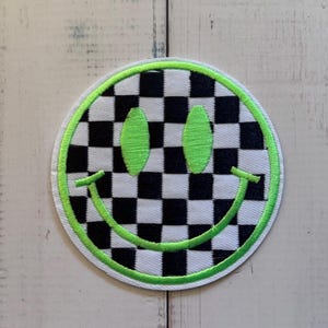 May include: A circular embroidered patch featuring a black and white chequered pattern. The design includes two oval-shaped eyes and a curved mouth, all stitched in a vibrant neon green thread. A neon green border outlines the entire circular shape.