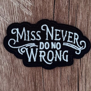 May include: A black embroidered patch with white stitched lettering that reads "MISS NEVER DO NO WRONG" in a stylised, cursive-inspired font. The patch has a scalloped, irregular border and is designed for application onto fabric surfaces.