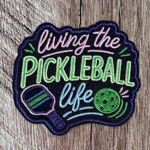 May include: A black embroidered patch featuring the text "living the PICKLEBALL life" in pink, light green, and light blue thread. The design includes a stylized pickleball paddle and a green perforated ball with motion lines.