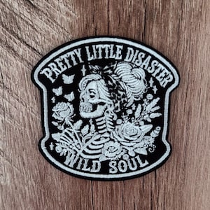 May include: A black and white embroidered patch featuring a skeleton with a hair bun and a patterned headband. The design includes roses, small butterflies, and the text PRETTY LITTLE DISASTER at the top and WILD SOUL at the bottom in white thread.