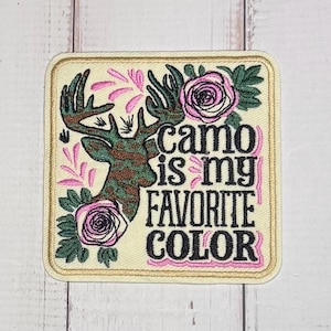 May include: A square embroidered patch with a cream background featuring a camouflage-patterned deer head, two pink roses with green leaves, and the black embroidered text "camo is my FAVORITE COLOR" with pink accents and a gold border.