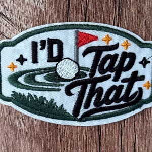 May include: An oval embroidered patch with a white background and a dark green border. It features a golf ball on a green, a red flag, and the black embroidered text I'D Tap That. Four small orange four-pointed stars are scattered around the design.
